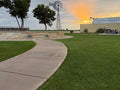 A curved concrete pathway cuts through a large Palisades Zoysia lawn toward a stone pool and trees, with a windmill and industrial-chic storage container against an orange sunset sky.