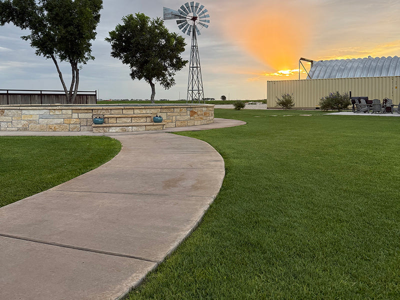 A curved concrete pathway cuts through a large Palisades Zoysia lawn toward a stone pool and trees, with a windmill and industrial-chic storage container against an orange sunset sky.