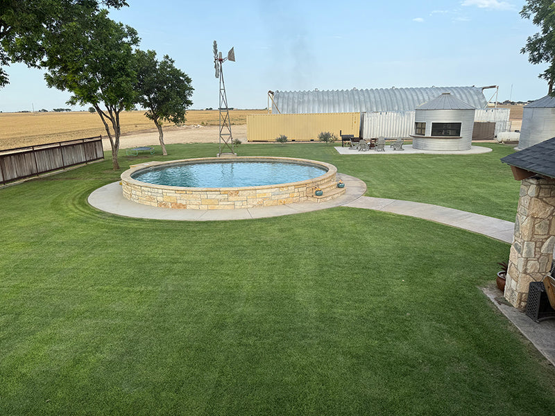 A sweeping scene showing an outdoor living space with a circular stone pool as a focal point, set on a luxurious Palisade Zoysia lawn. Trees, a windmill, and a silo converted to a kitchen and bar are visible in the background under a blue sky.