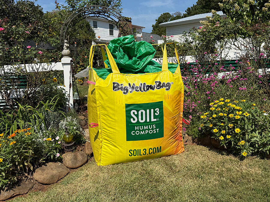 Yellow SOIL3 Humus Compost bag in a garden setting with flowers and plants.